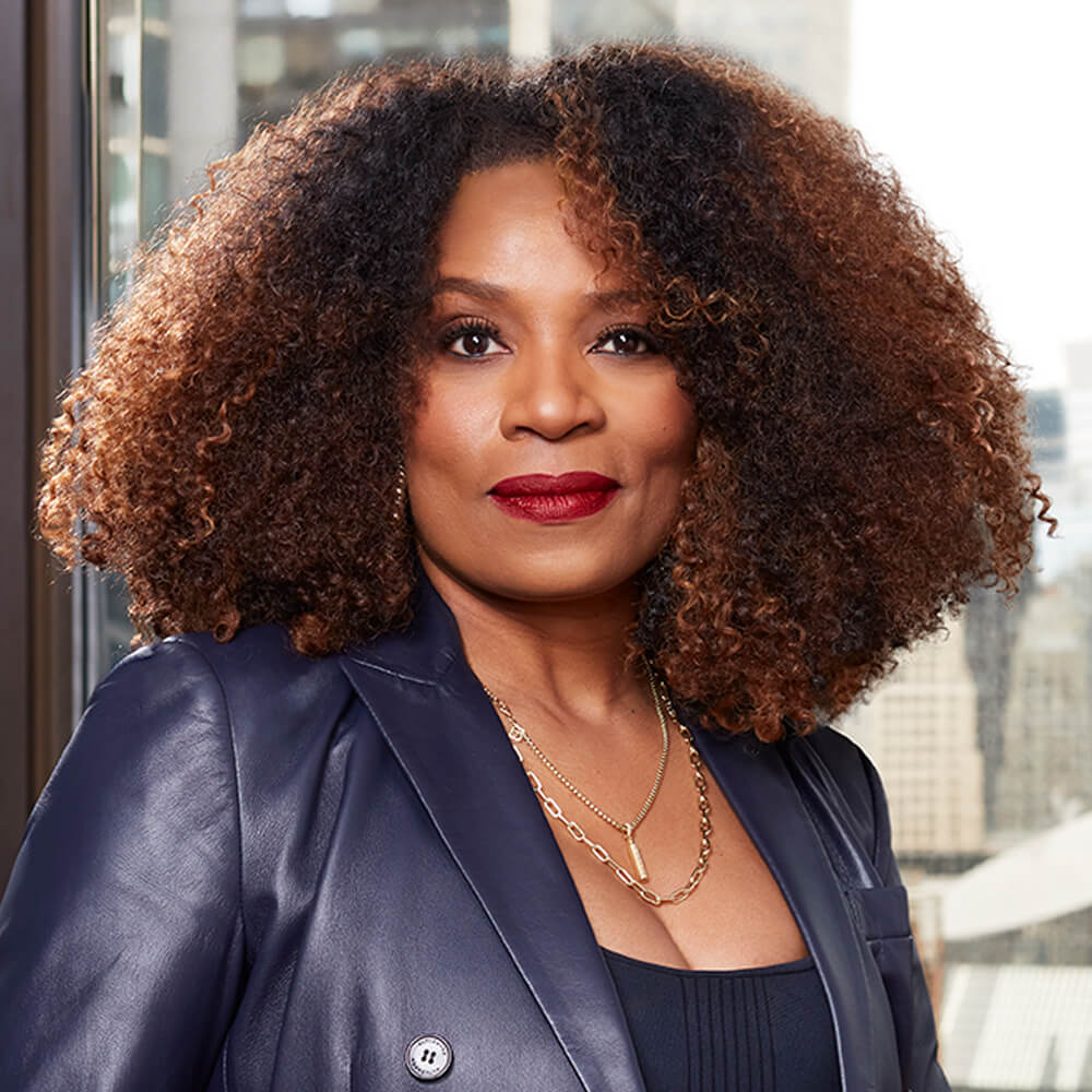 A woman with curly hair and red lipstick wearing a dark blazer and layered necklaces stands in front of a window with city buildings in the background.