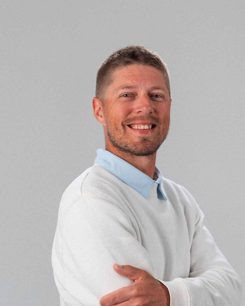 Derrick Ottenbreit, a man with short brown hair and a beard, wearing a white sweater over a light blue collared shirt, stands smiling with arms crossed against a plain light gray background.