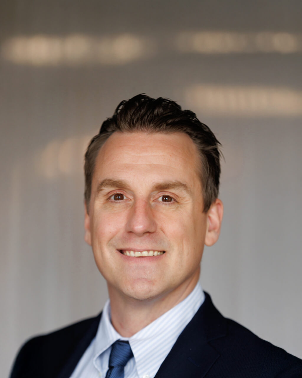 Eric J. Korslin, dressed in a dark suit, light blue shirt, and tie, smiles at the camera against a neutral, softly blurred background.