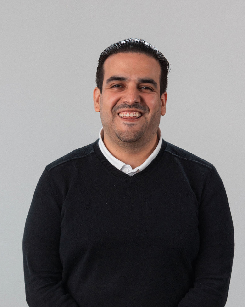 Fabrithzio, a man with dark hair wearing a black sweater over a white collared shirt, stands against a plain light background and smiles at the camera.