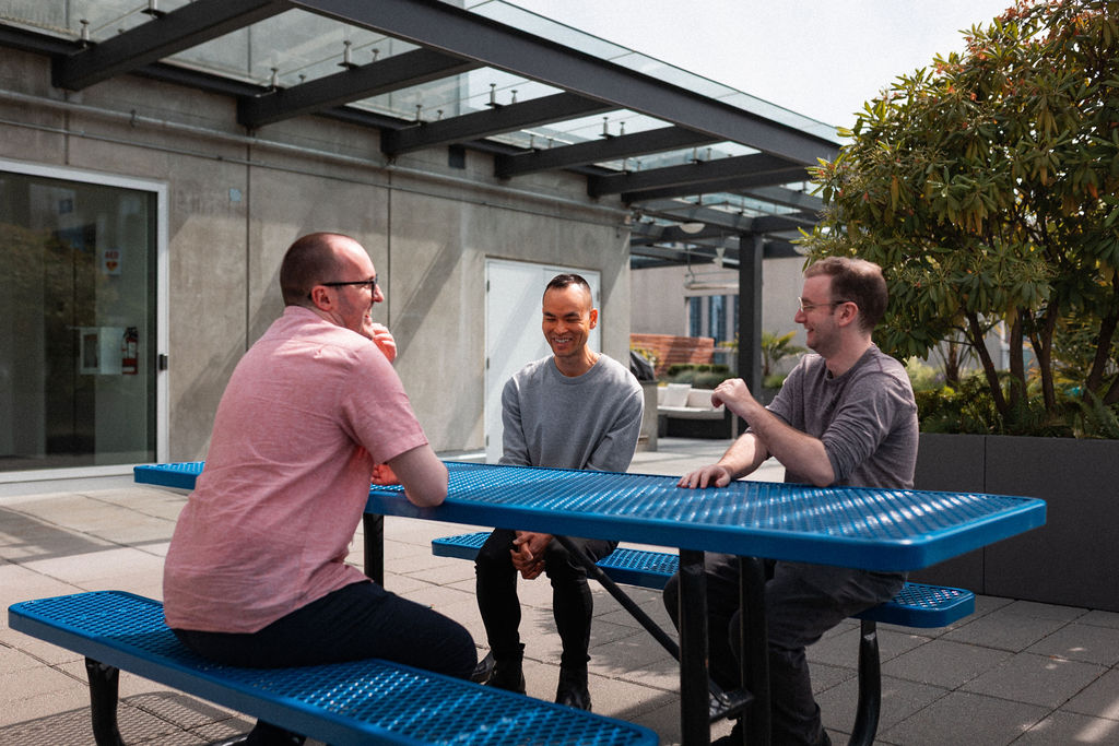 Three men sit and talk at a blue picnic table on an outdoor patio near a modern building with glass panels and potted plants.