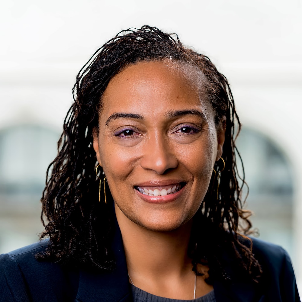 A woman with medium-length curly hair, wearing a dark blazer and earrings, smiles at the camera against a blurred light background.