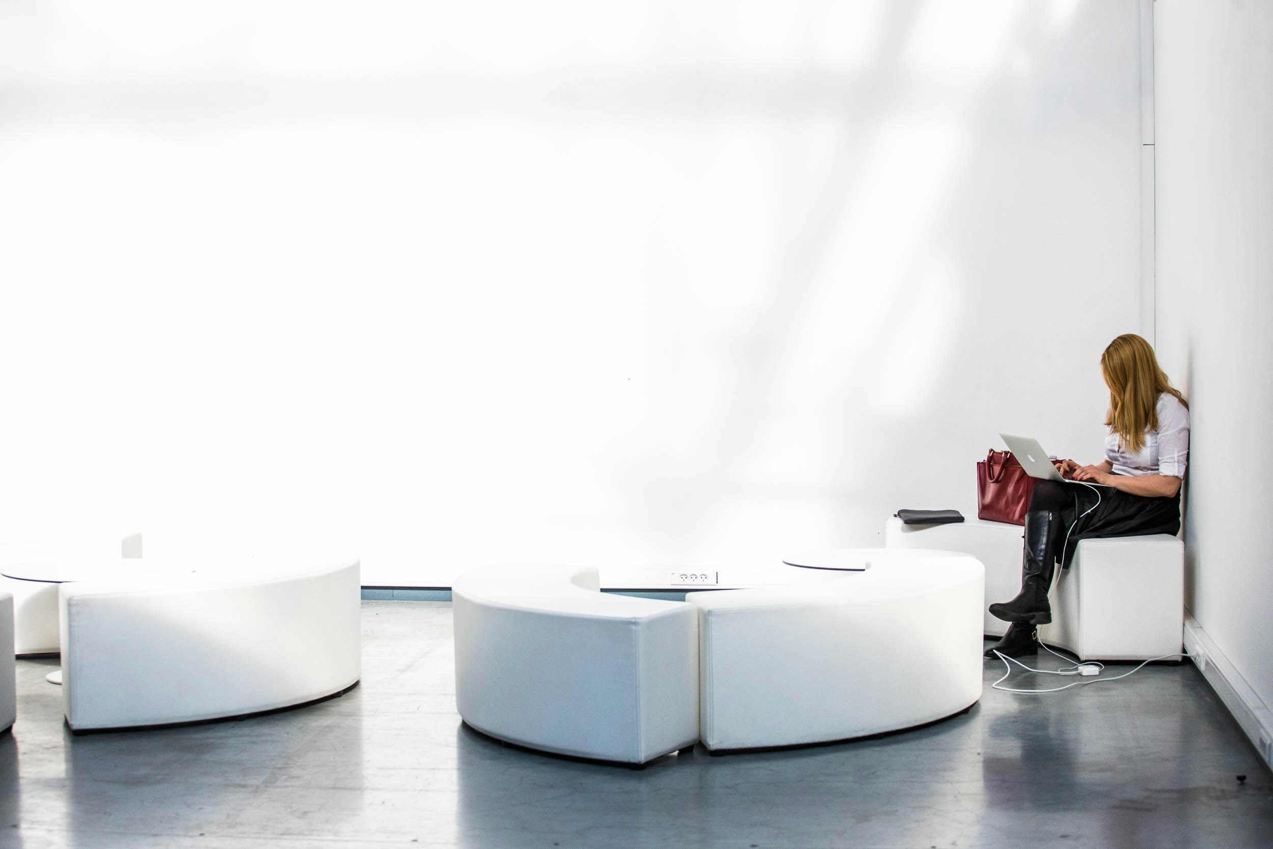 Una mujer está sentada sola en un banco blanco curvado, trabajando con un ordenador portátil y con un bolso rojo a su lado, en una habitación luminosa y minimalista.