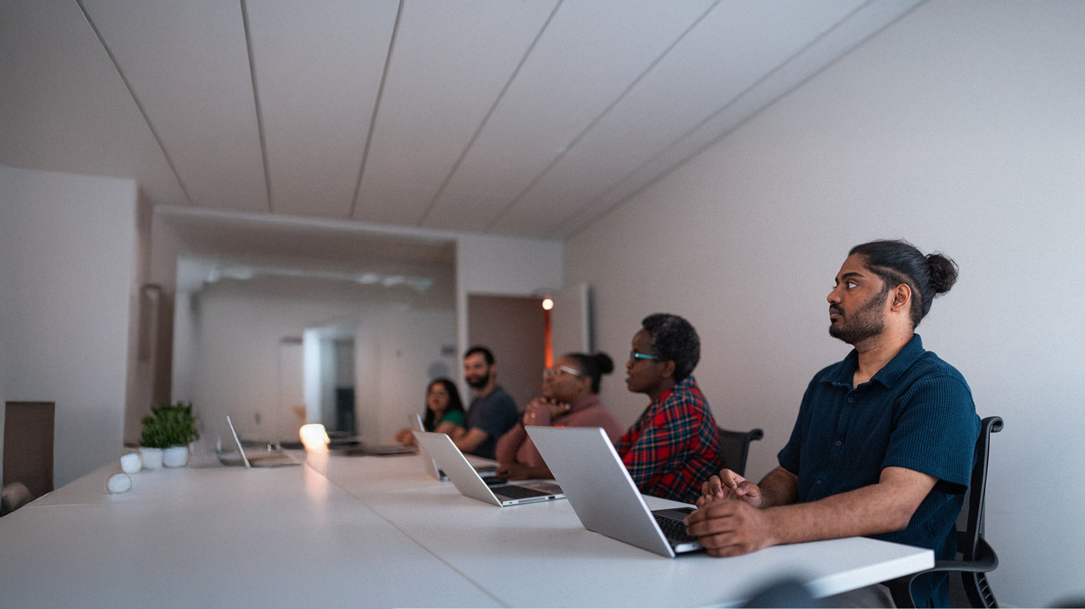 Five people sit at a conference table with laptops open, facing forward and appearing attentive during a meeting in a modern, well-lit office space.