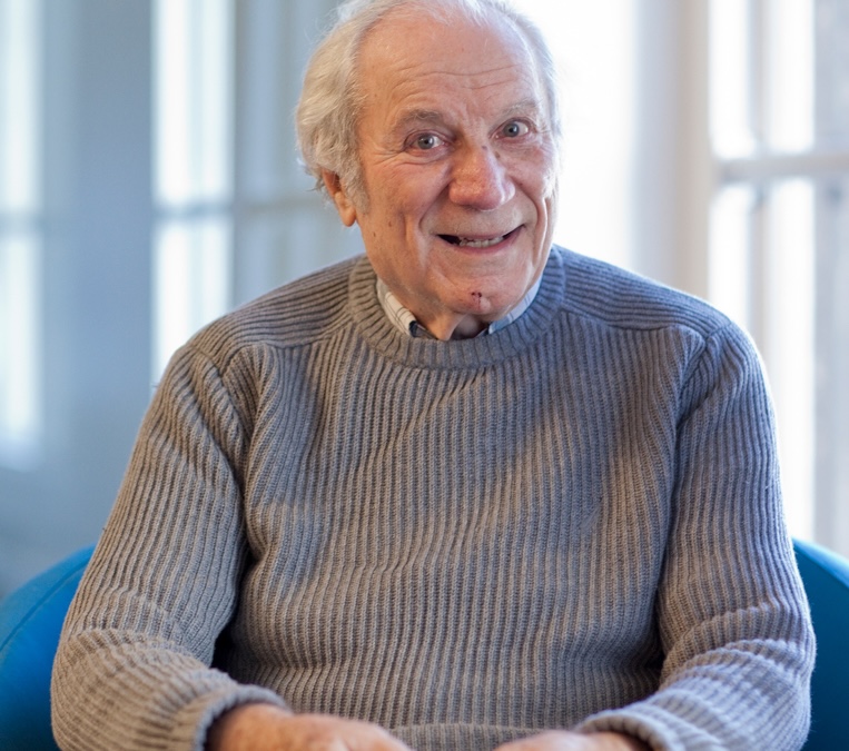 An elderly man with gray hair, wearing a ribbed gray sweater, sits and smiles at the camera in a bright indoor setting.