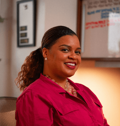Lunden Gregory, a woman with curly hair pulled back, wears a bright pink top and a beaded necklace as she sits and smiles in an office setting with a whiteboard and framed items in the background.