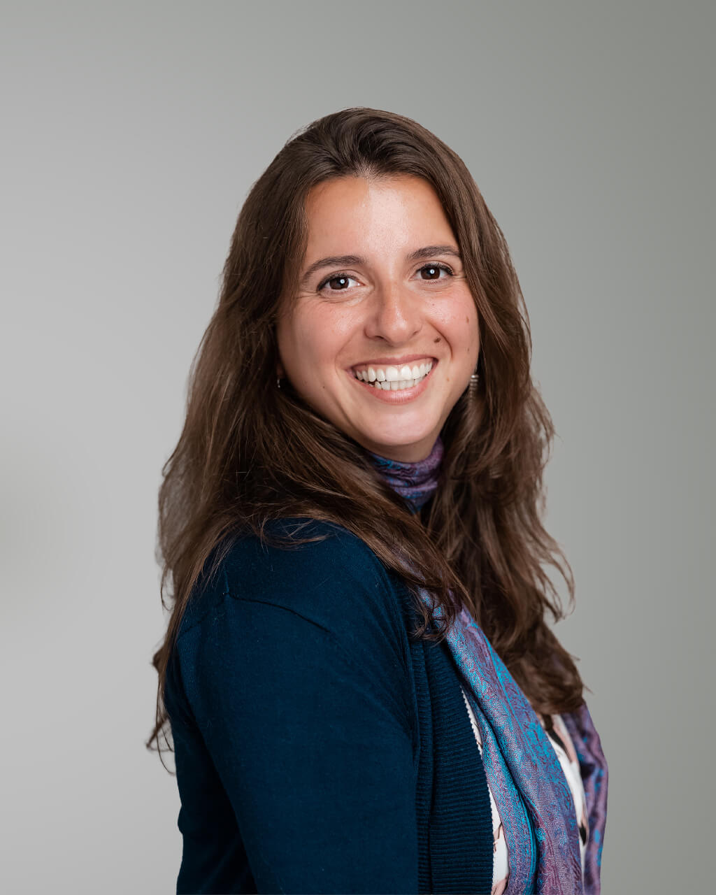 Ioana Birleanu, a woman with long brown hair, smiles warmly while wearing a dark cardigan and patterned scarf, posing against a plain light grey background.