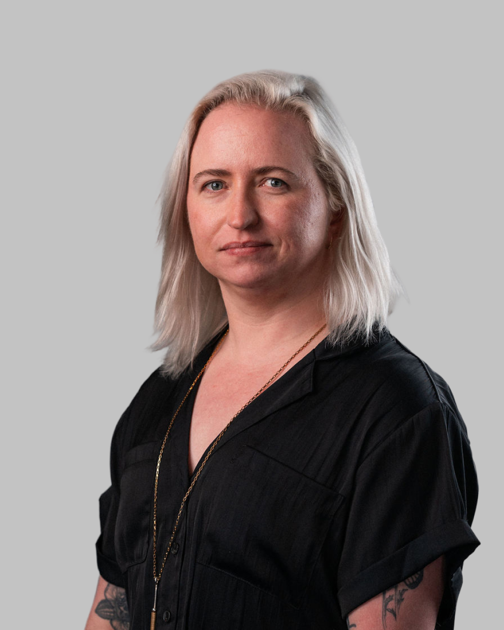 A person with shoulder-length blonde hair and tattoos, wearing a black shirt and necklace, poses in front of a plain gray background—effortlessly channeling the cool confidence of Jenii.