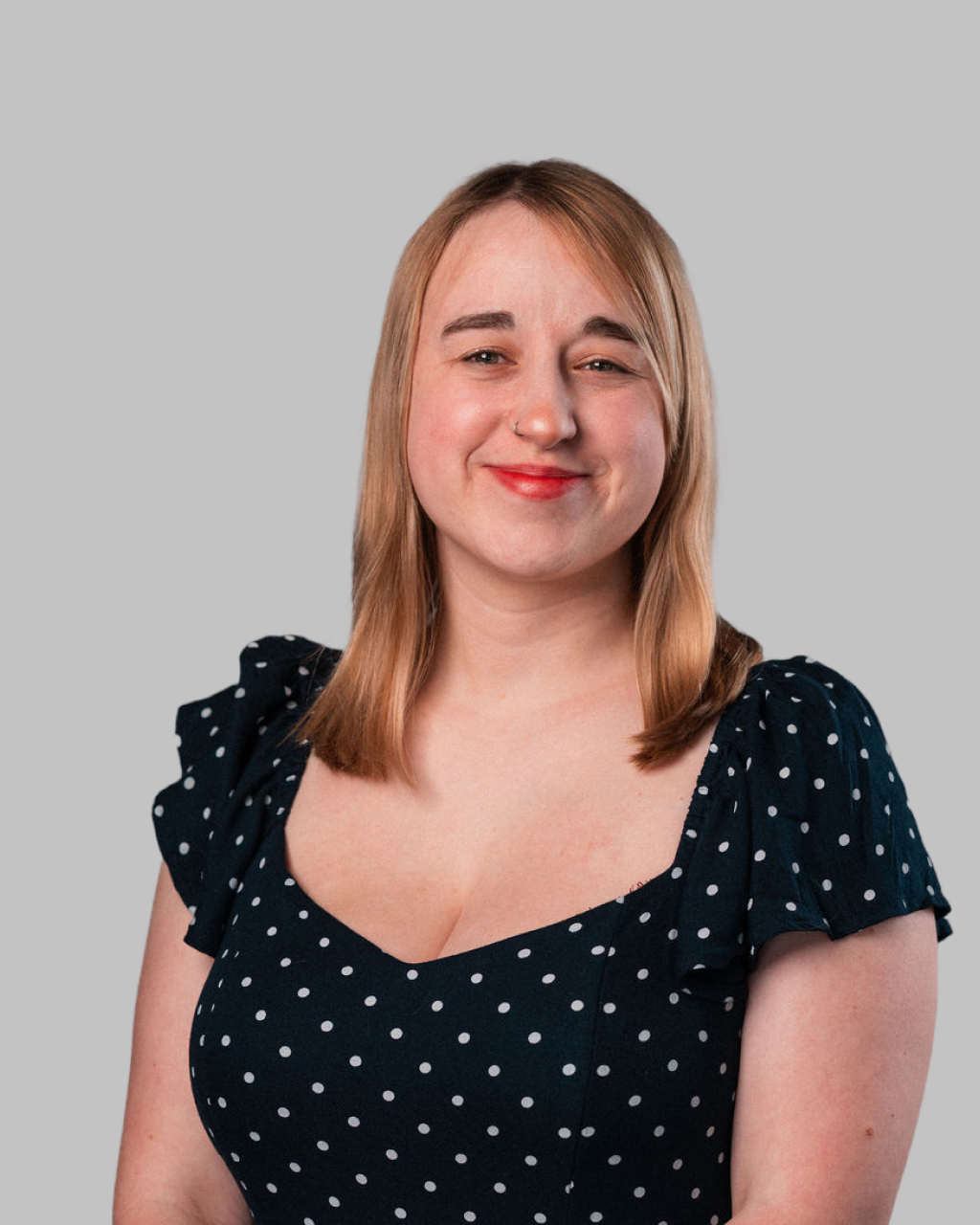 Jenny Michalski, with straight blonde hair, smiles in a black dress with white polka dots against a plain grey background.