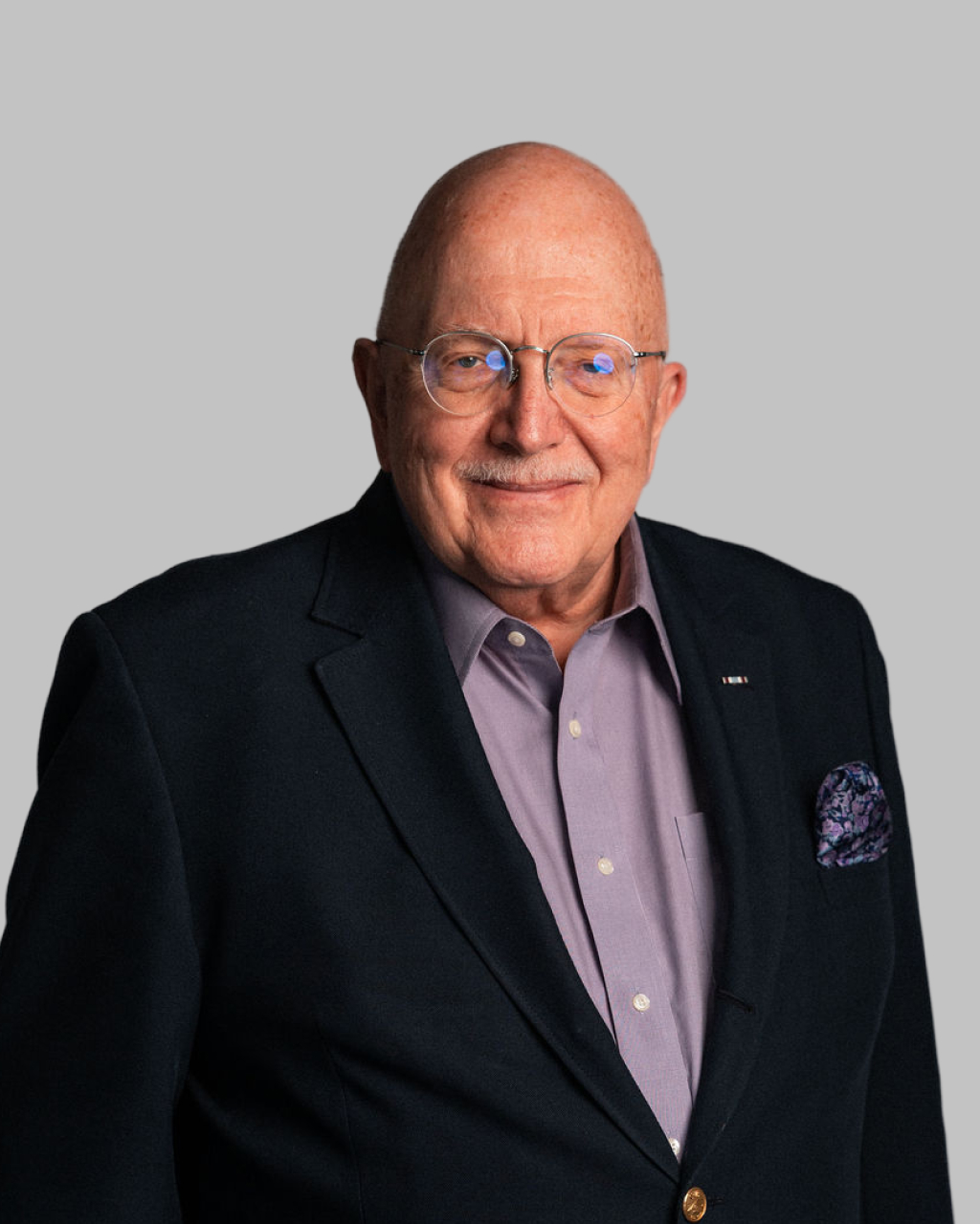 Joseph E. Troiani, an older man with glasses, wearing a dark blazer over a light purple shirt, stands against a plain gray background.