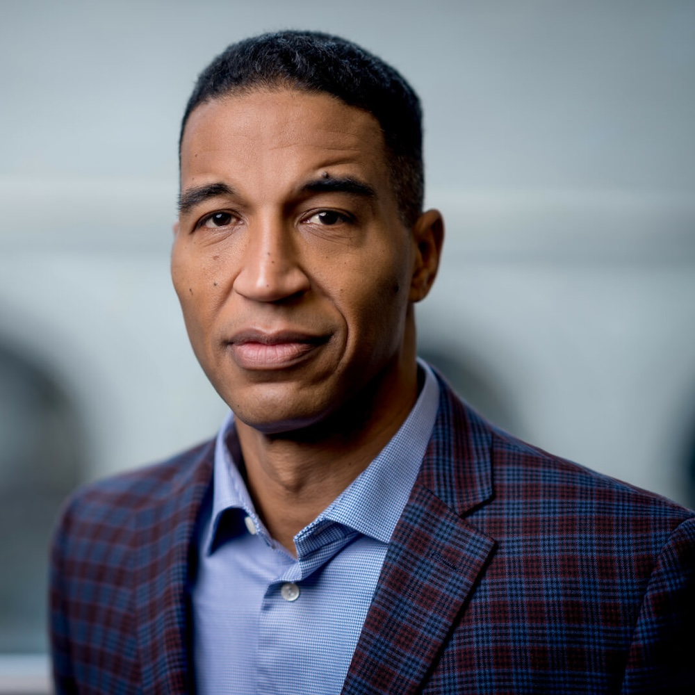 A man in a checked blazer and blue shirt stands indoors, facing the camera with a neutral expression. The background is softly blurred.
