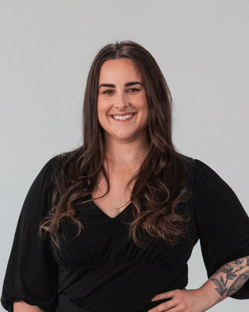 Kelsey Sarasqueta-Allen, a woman with long brown hair wearing a black top, stands against a light gray background, smiling with one hand on her hip.