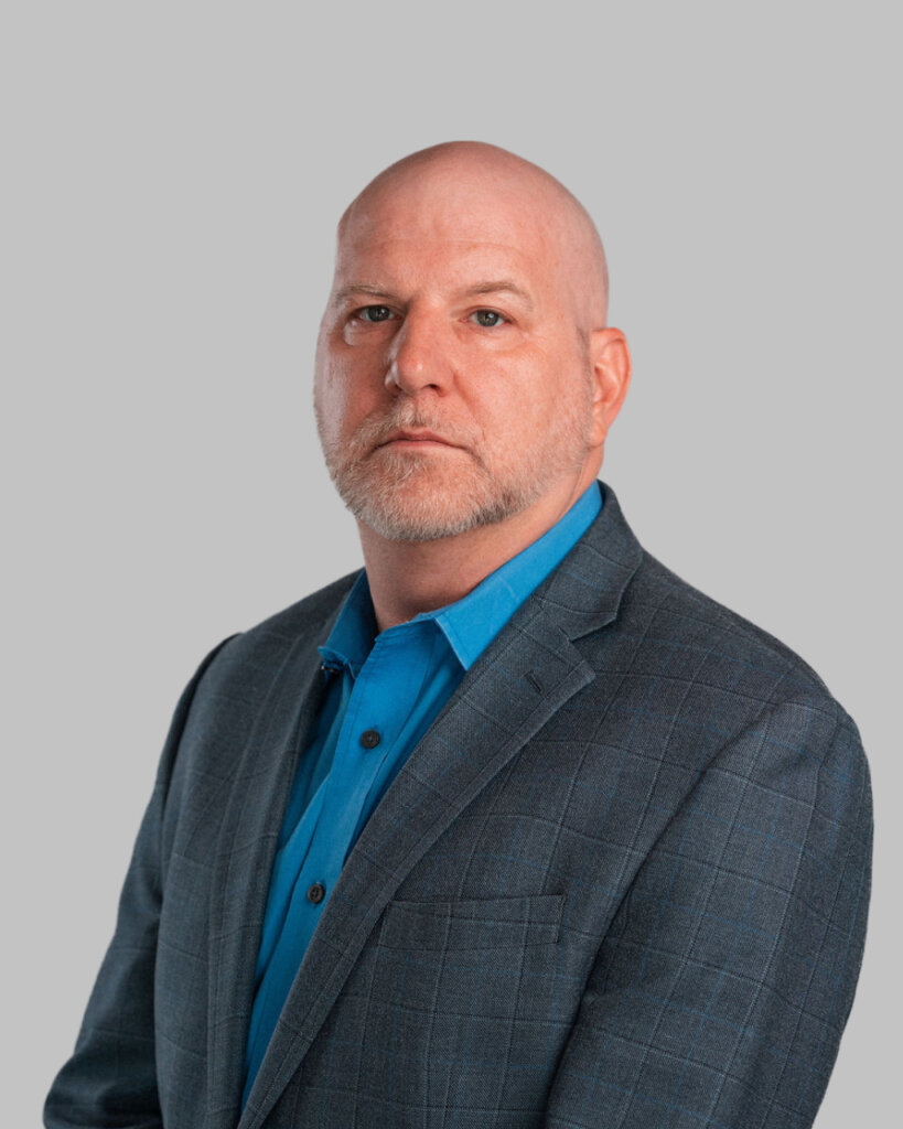 Kevin A. Osten-Garner, a bald man with a trimmed beard, wears a blue dress shirt and plaid blazer while posing against a plain gray background.