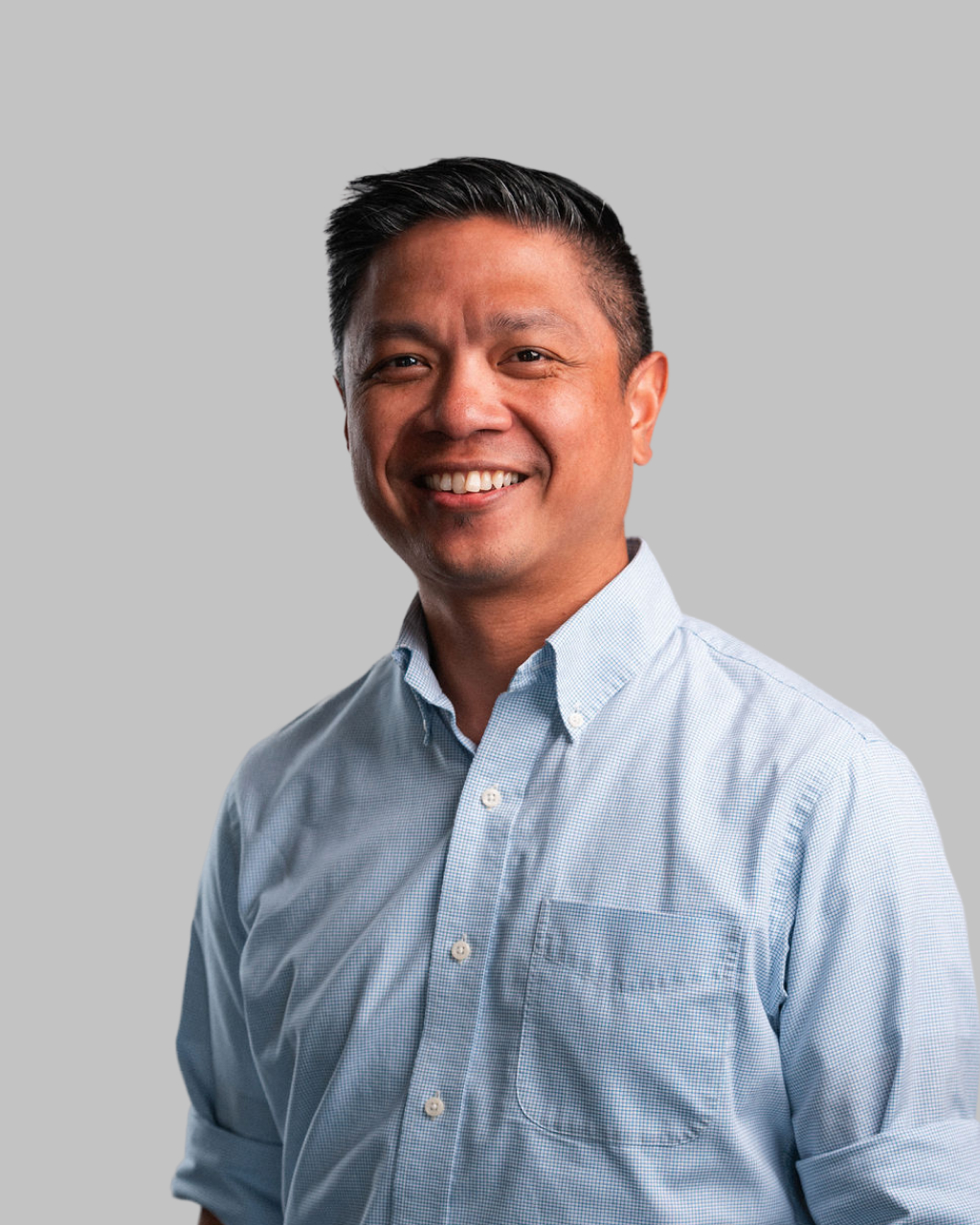A man wearing a light blue button-down shirt smiles while standing against a plain gray background in a portrait by Kimber Solana.