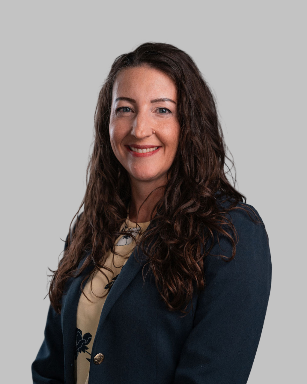 Woman with long brown wavy hair wearing a navy blazer and beige top, smiling, posed against a plain light gray background.