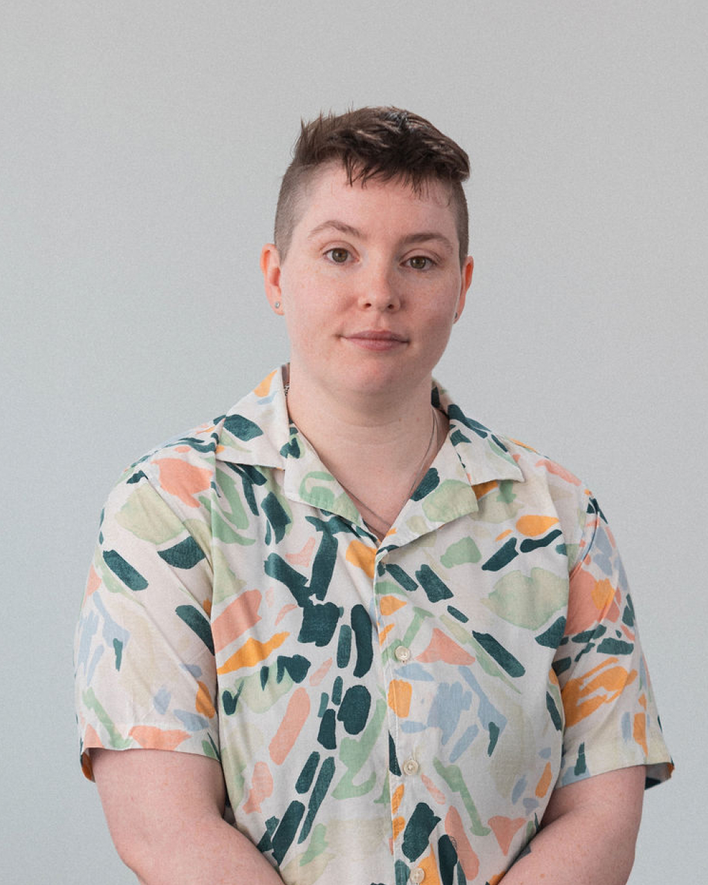 Lauren, with short hair and a patterned short-sleeve shirt, stands against a plain light-colored background, looking at the camera.