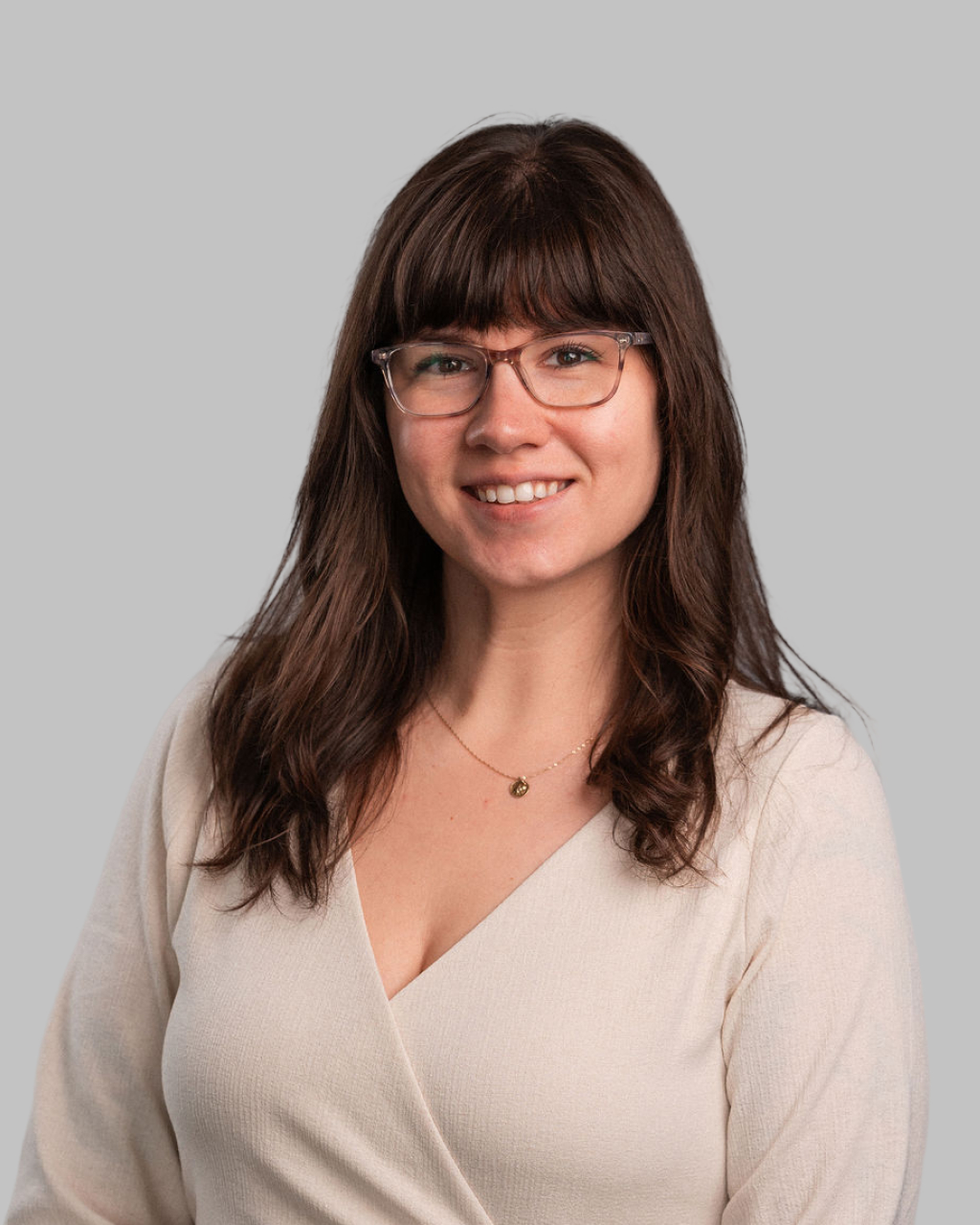 Liz Blondel, a woman with long brown hair and glasses wearing a cream-colored wrap top, smiles at the camera against a plain gray background.