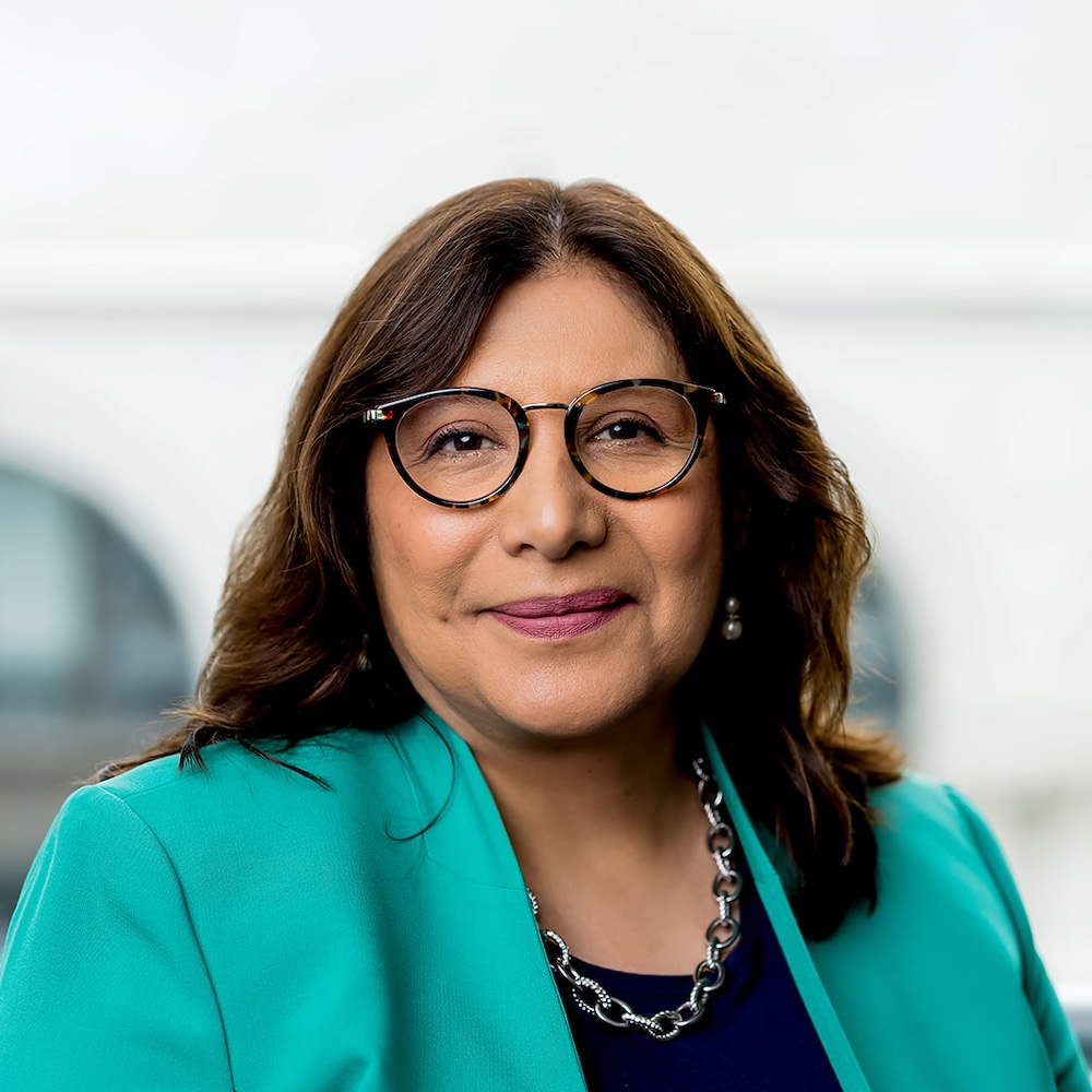 A woman with long brown hair and glasses, wearing a turquoise blazer, navy top, and silver necklace, poses in front of a blurred background.