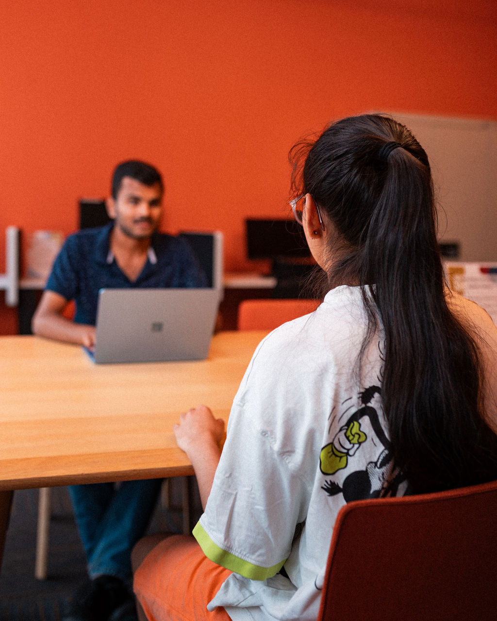 Two people sit across from each other at a table in an office, with one facing a laptop and the other facing away from the camera.
