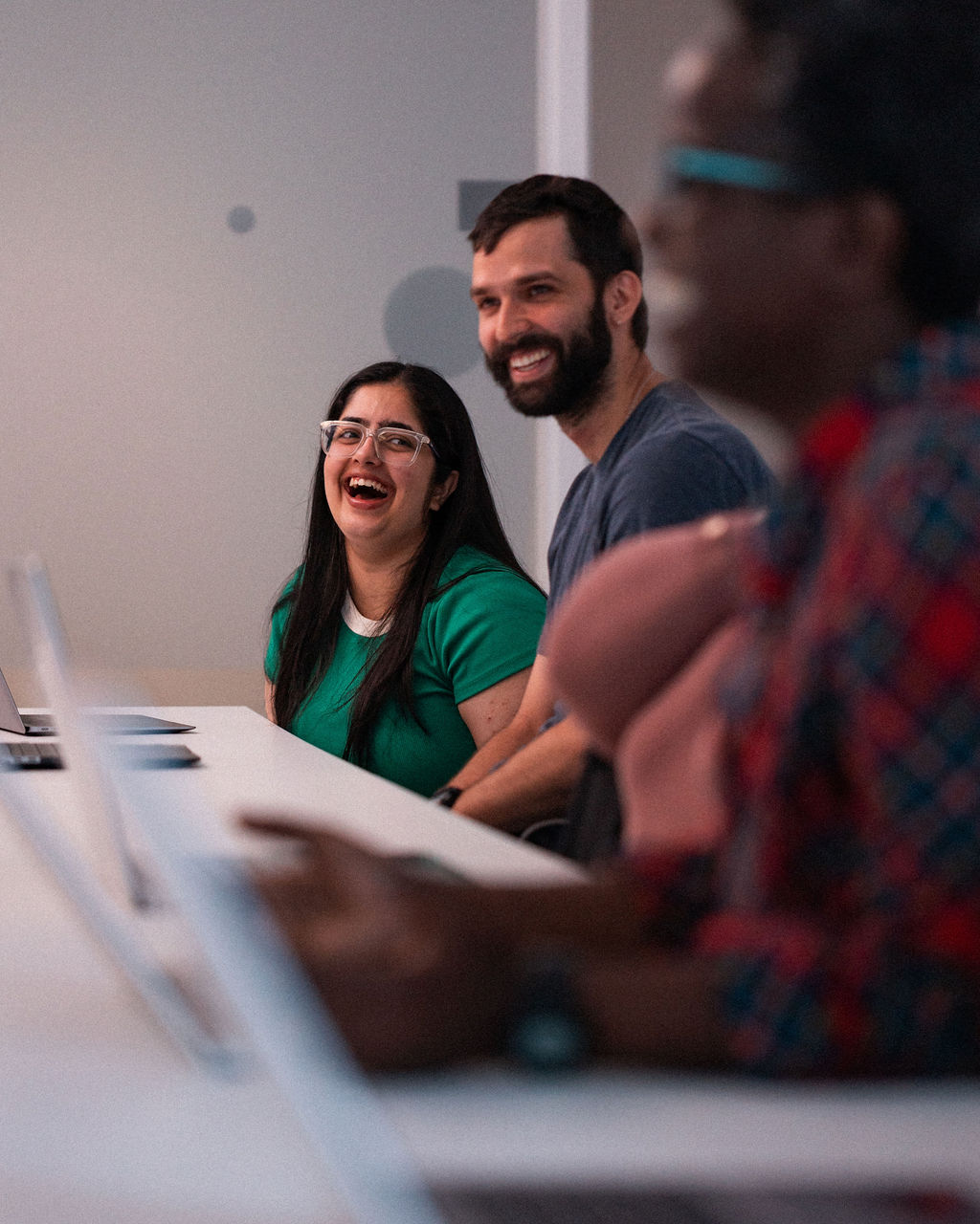 Three people sitting at a table with laptops, two of them smiling and laughing, in a brightly lit indoor setting.