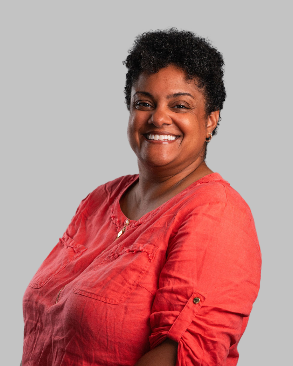 Michelle Anderson, a woman with short curly hair, wearing a coral-colored shirt, stands smiling against a plain gray background.