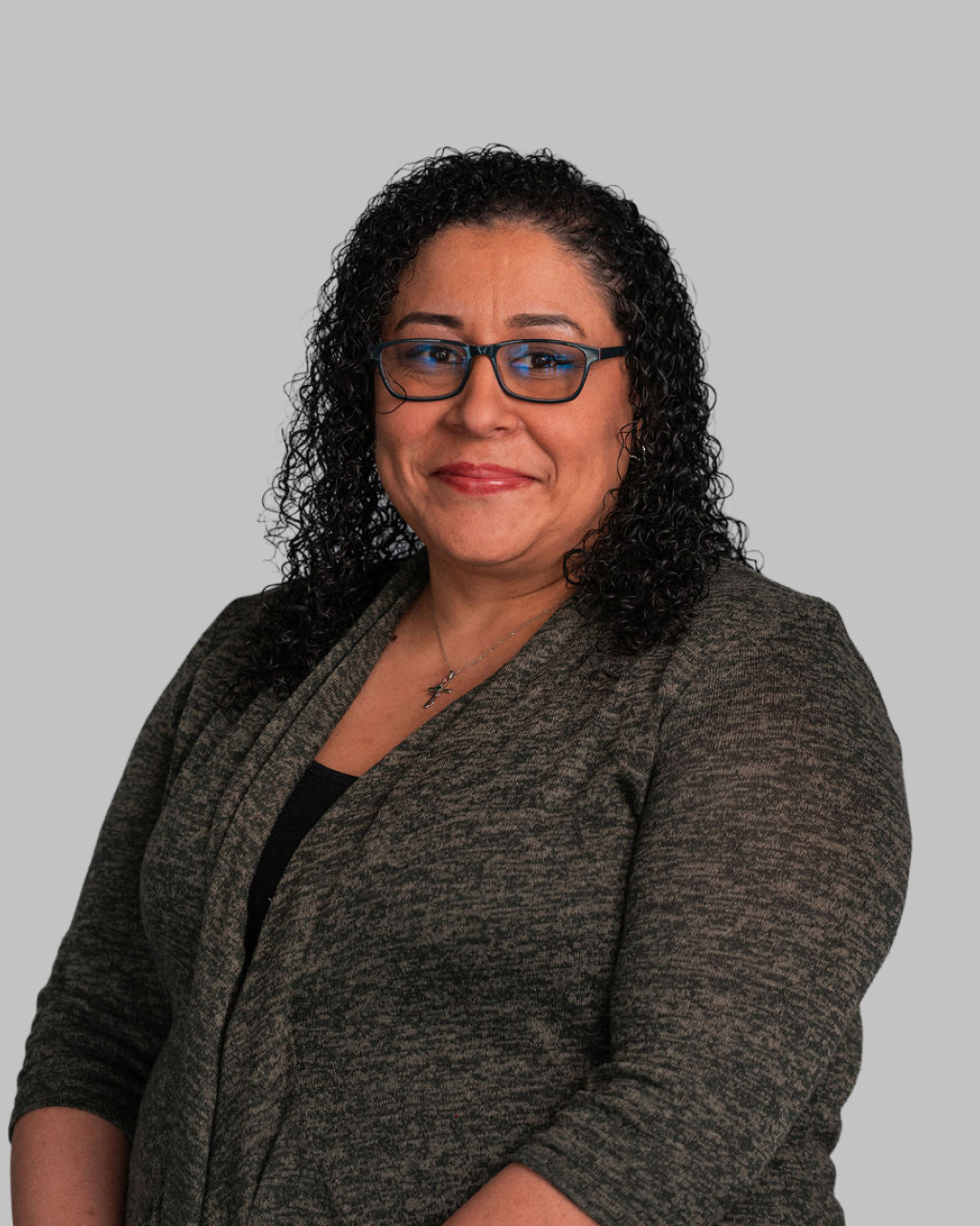 Michelle Tiwade, a woman with curly dark hair, wearing glasses and a grey sweater, smiles at the camera against a plain grey background.