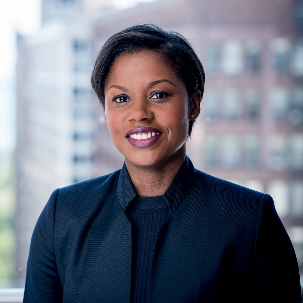 A woman with short dark hair in a dark blazer smiles in front of a window with blurred city buildings in the background.