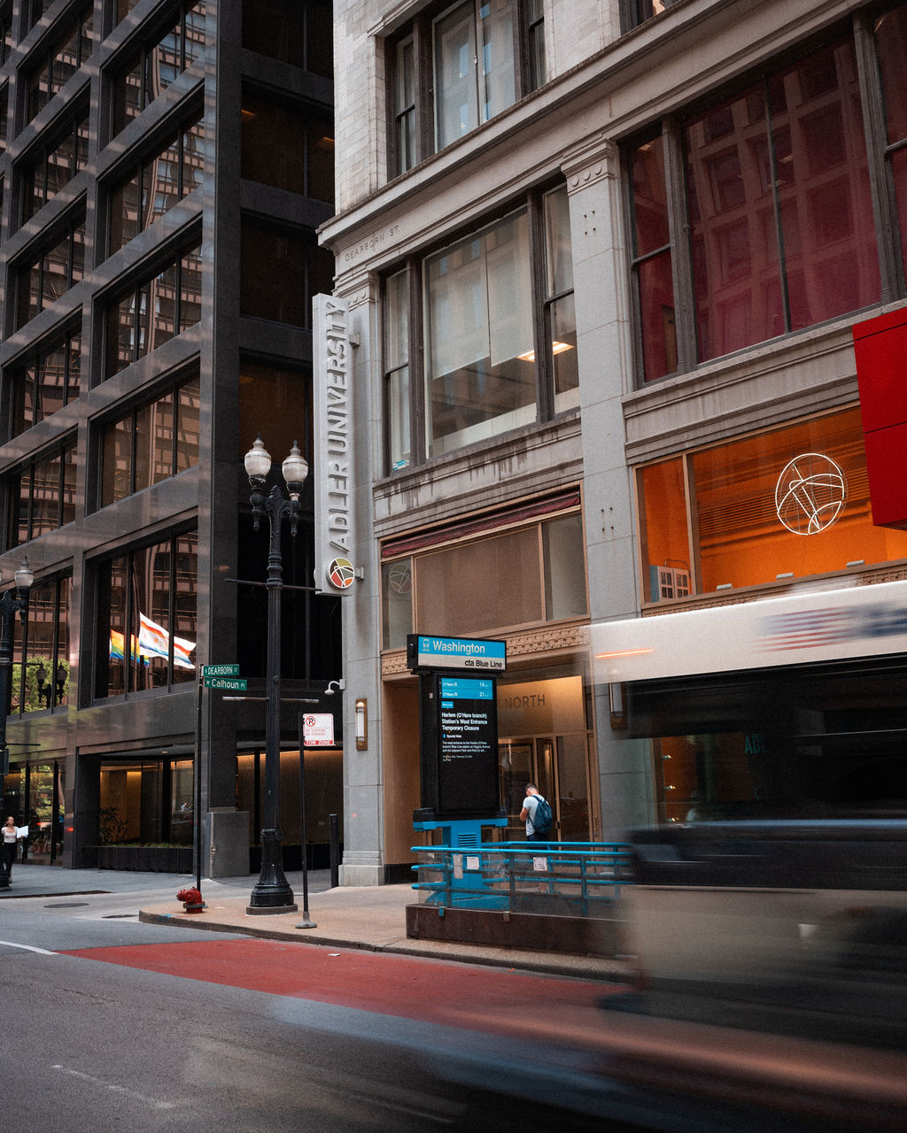 A city street with a bus passing by, a person waiting at a bus stop, and tall office buildings in the background.