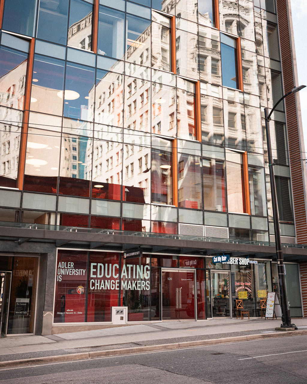 The glass facade of Adler University reflects nearby buildings. Signs read "EDUCATING CHANGE MAKERS" and "Barber Shop" above the street-level entrances.