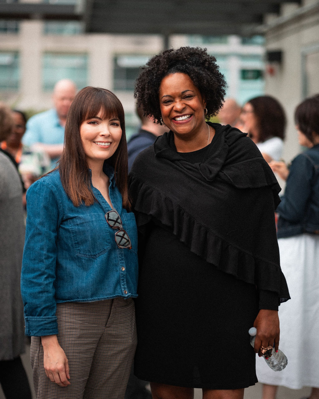 Two women stand side by side, smiling at an outdoor social gathering that celebrates shared values, with several people blurred in the background.