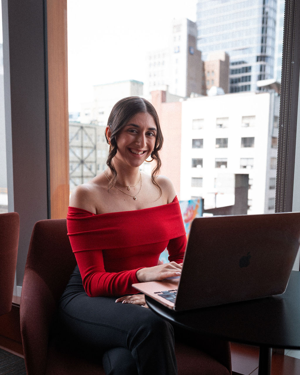 A woman in a red off-shoulder top sits at a table with a laptop, smiling, with city buildings visible through the window behind her.