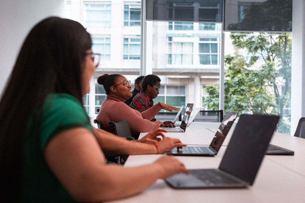 Three people sit at a table with laptops in a modern office, working and discussing while facing large windows that show an urban building outside.