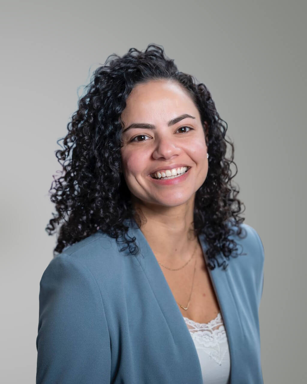 Reylla Santos, a woman with curly dark hair, wears a blue blazer and white top, smiling at the camera against a plain light background.