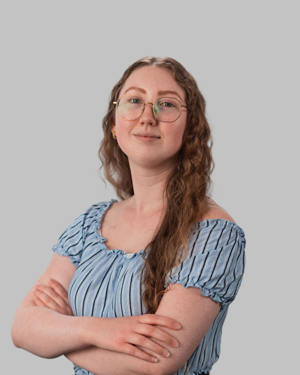 Rhiannon Fancett, a young woman with long wavy hair and glasses, stands against a plain gray background, wearing a blue striped blouse and crossing her arms.