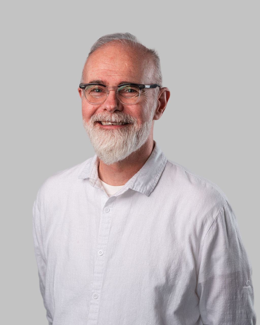 A middle-aged man with glasses, white hair, and a beard, wearing a white shirt, smiling in front of a plain grey background—Richard A. Niolon exudes warmth and approachability.