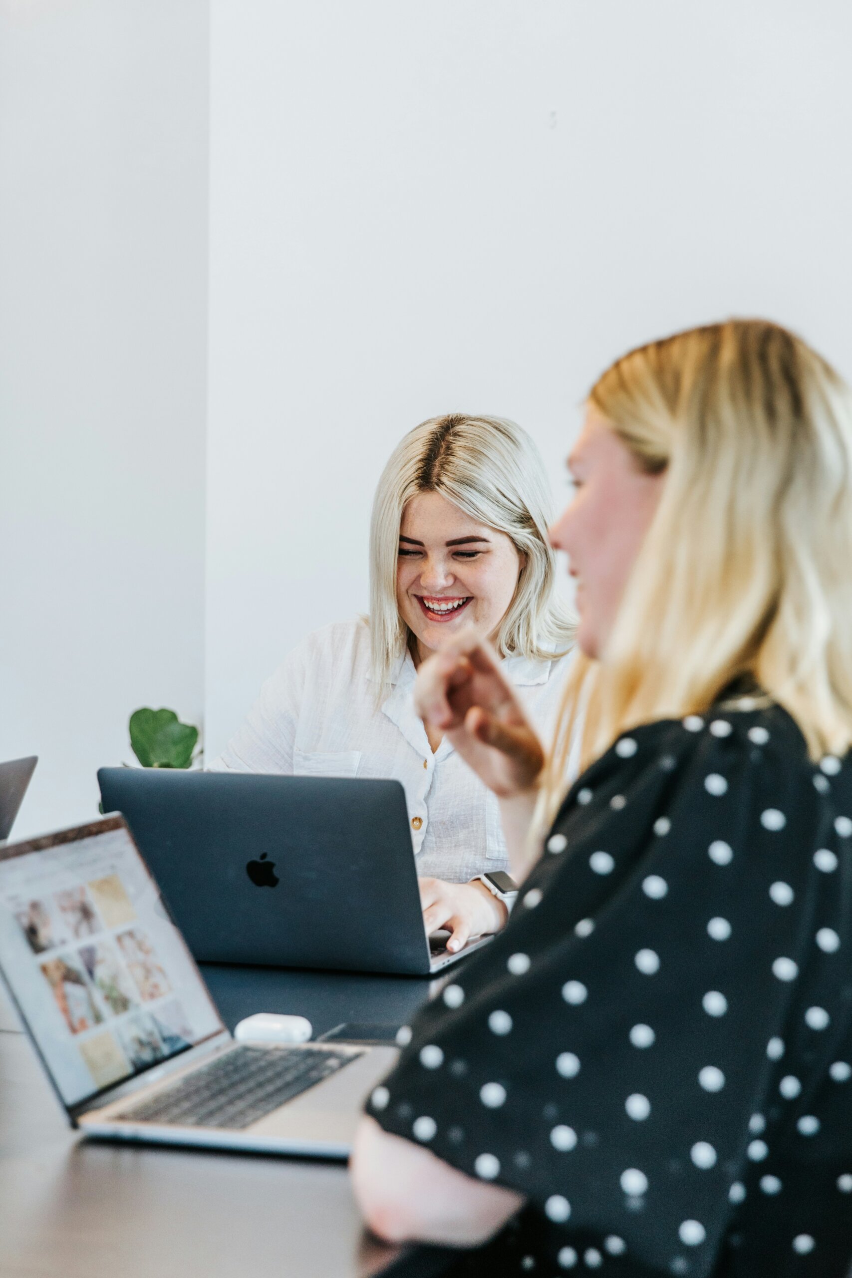 Two women with laptops sit at a table in a bright room, one smiling while looking at her screen and the other gesturing with her hand.