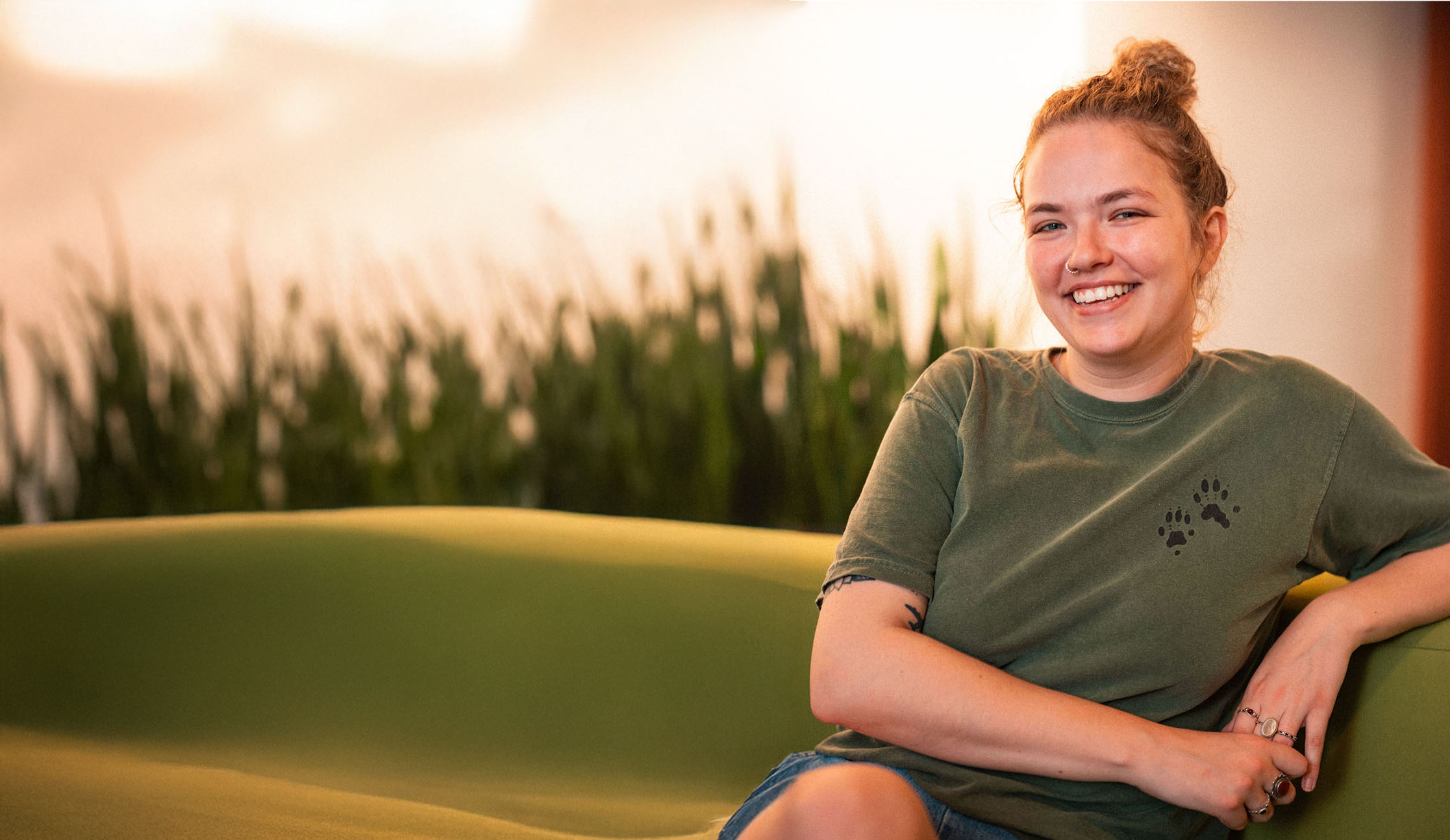 An international student with a bun hairstyle sits on a green sofa, smiling at the camera, wearing a green t-shirt and denim shorts, with plants in the background.
