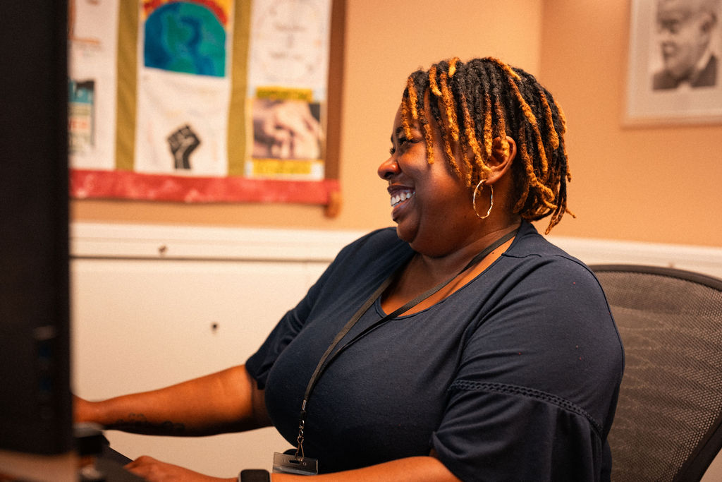 Una mujer con el pelo corto y rizado está sentada ante un escritorio, sonriendo mientras mira la pantalla del ordenador; en la pared que hay detrás de ella se ven unas ilustraciones de colores.