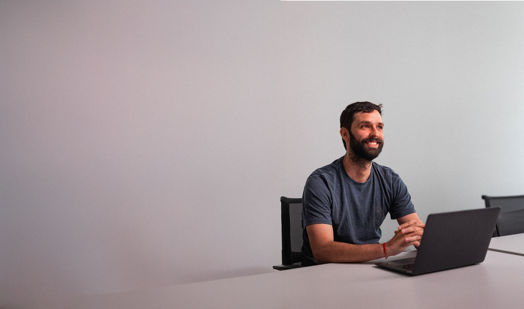 A man with a beard sits at a table in front of a laptop, smiling, in a minimalist office with a plain light-colored wall.