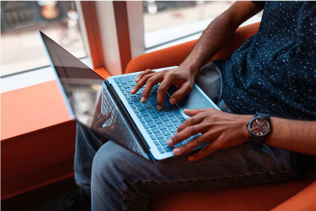 A person sits on an orange chair by a window, typing on a blue laptop resting on their lap.