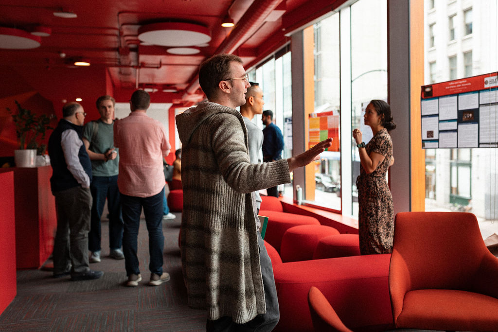 People stand and converse in a modern, red-walled lounge with large windows and posters displayed on the glass.