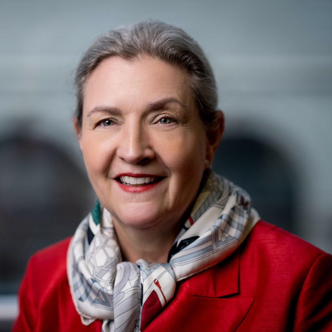 A woman with gray hair wearing a red blazer and a patterned scarf, smiling at the camera against a blurred background.