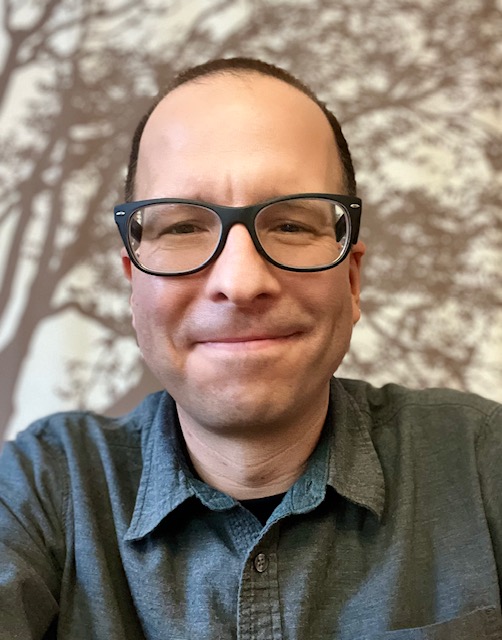 Jason Grebasch, wearing glasses and a button-up shirt, smiles at the camera with a tree-patterned background behind him.