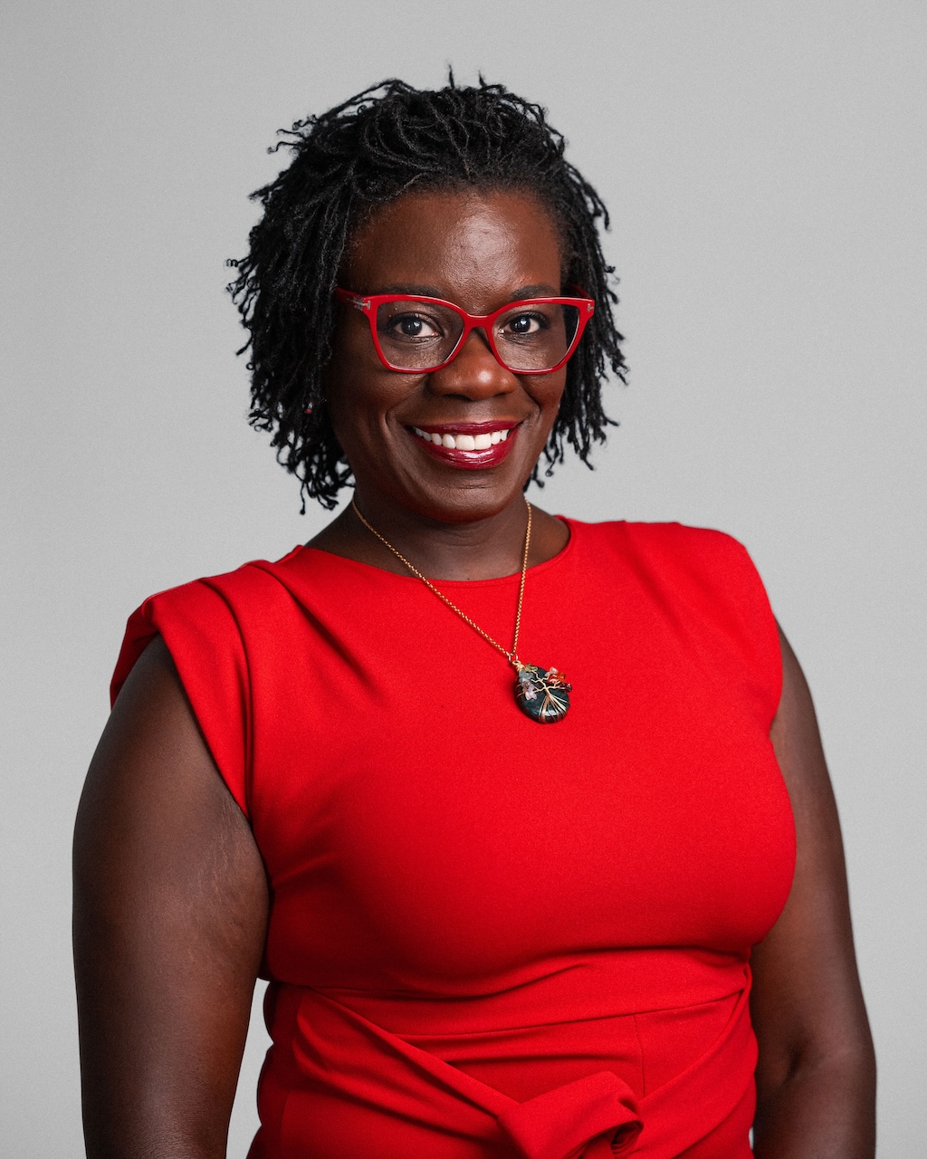 A woman with short braided hair, wearing red glasses, a red sleeveless dress, and a pendant necklace, smiles at the camera against a plain light gray background.