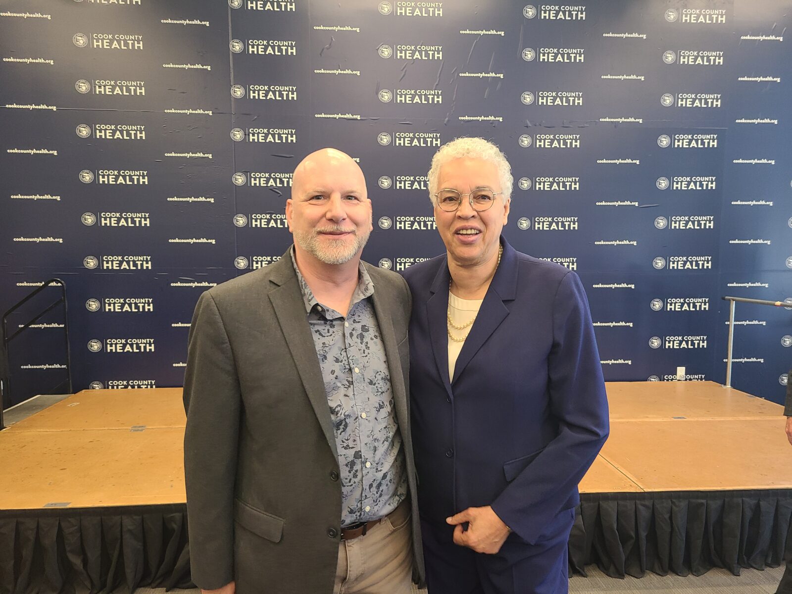 Two people stand side by side, smiling, in front of a Cook County Health step-and-repeat backdrop at an indoor event celebrating the Behavioral Health Grant.