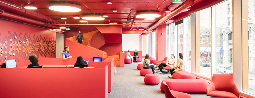 A modern office space with red walls and furniture, large windows, people working at desks, and others sitting or talking in lounge chairs.