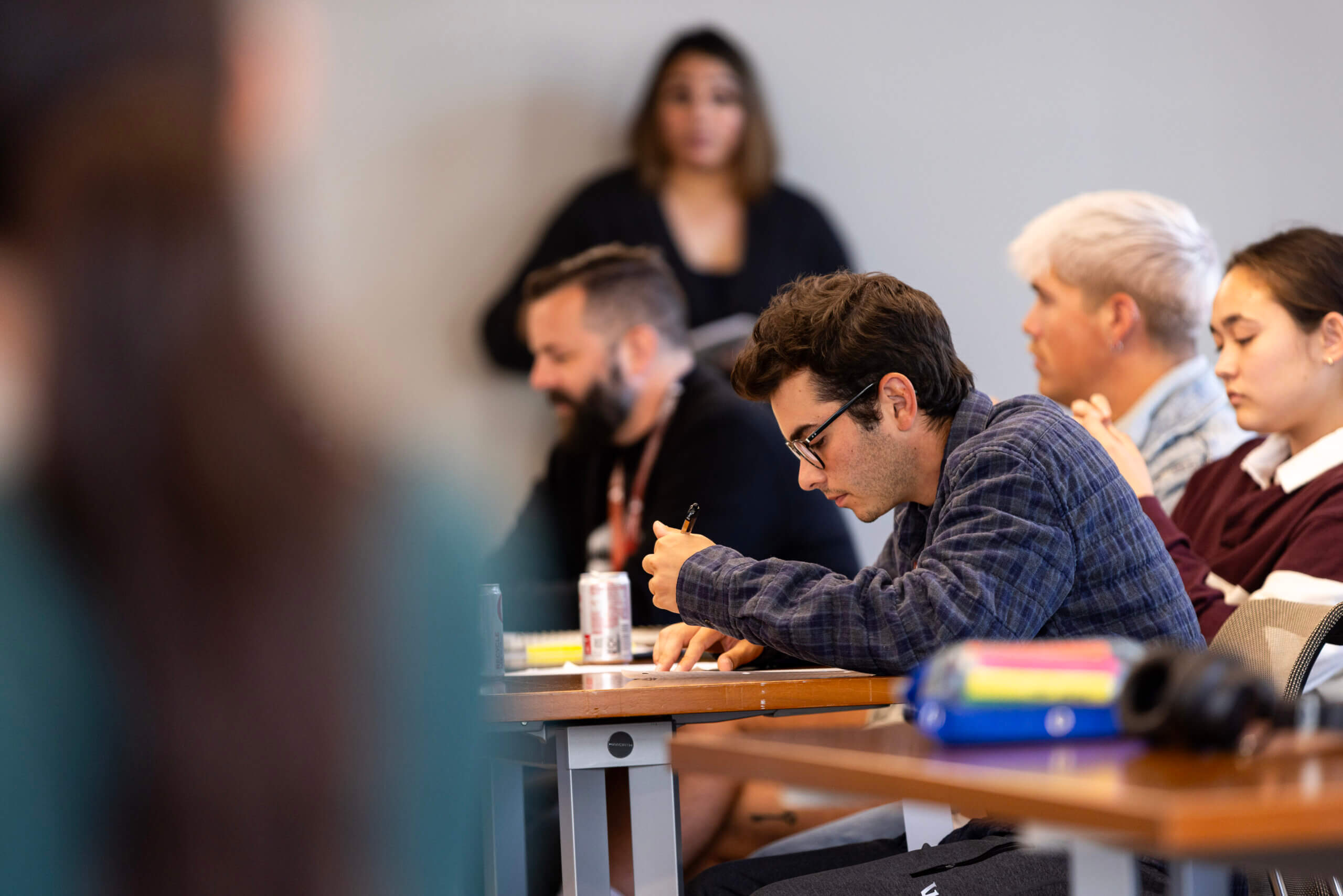 A group of people sit at desks in a Clinical Mental Health Counseling, M.A. classroom, with one person in the foreground writing on paper and others listening or observing.