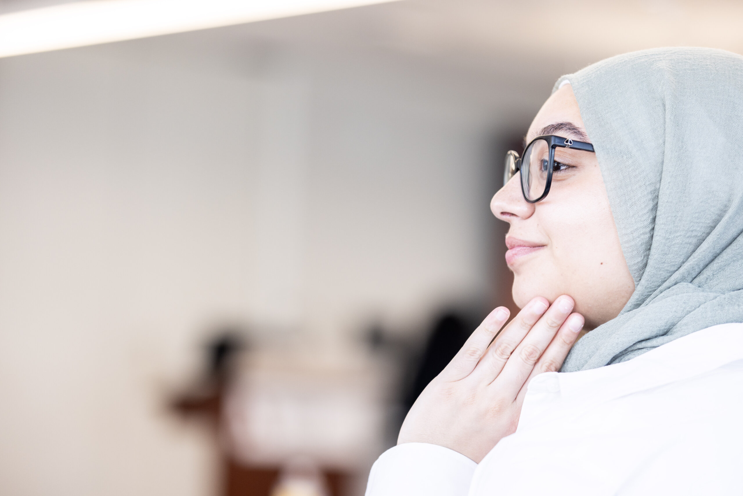 A woman wearing glasses and a light green hijab—perhaps a Clinical Mental Health Counseling, M.A. student—touches her neck and looks to the side, set against a blurred indoor background.