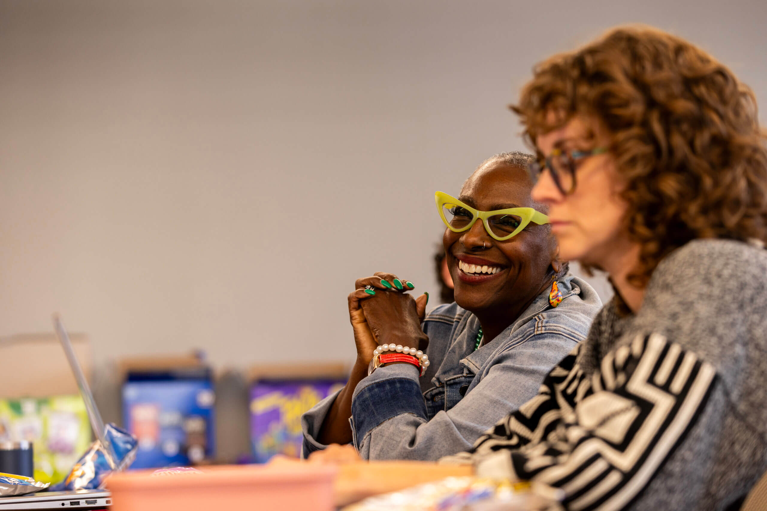 Two women sit at a table; one, possibly an MA in Clinical Mental Health Counseling student, is smiling and wearing green glasses, while the other is looking at a laptop.