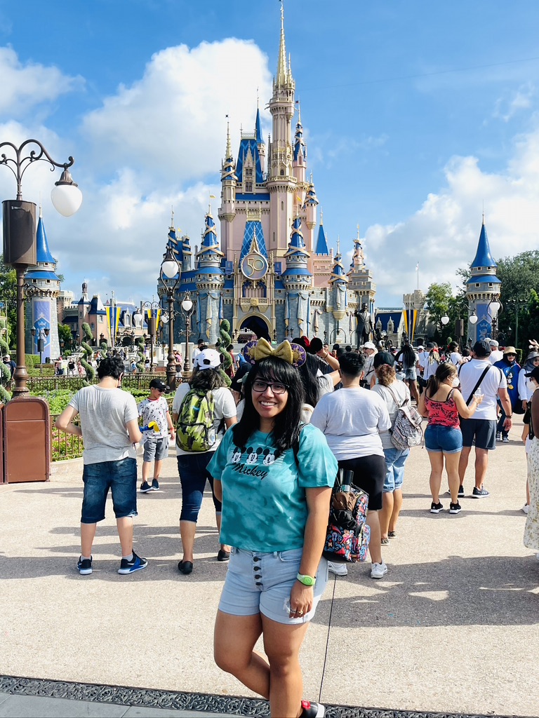 A Psy.D. student in a teal shirt and Minnie Mouse ears stands smiling in front of Cinderella Castle at a crowded Disney park on a sunny day.